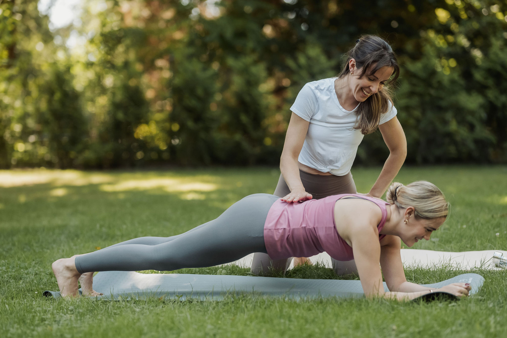 Zwei Frauen beim Training im Park, eine in der Plank-Position, die andere unterstützt sie.